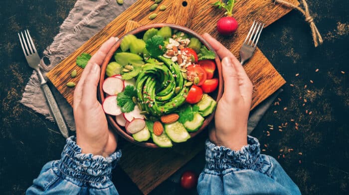 Healthy vegan salad, avocado, celery, cucumber, tomato, radish, nuts and seeds. Girl in denim blouse holding a bowl of vegan salad with visible hands.