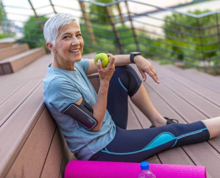 Sportos nő almát eszik. Ősz hajú nő a hatvanas évei elején sportedzés után pihen.