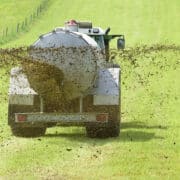 Egy traktor hátulról fotózva, amint trágyát szór szét a zöld, füves mezőgazdasági területen