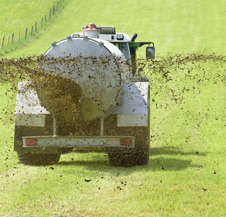 Egy traktor hátulról fotózva, amint trágyát szór szét a zöld, füves mezőgazdasági területen