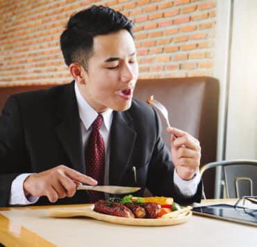 Egy öltönyös férfi ül egy étteremben, a villáján egy darab hús, a tányéron előtte hús és zöldségek