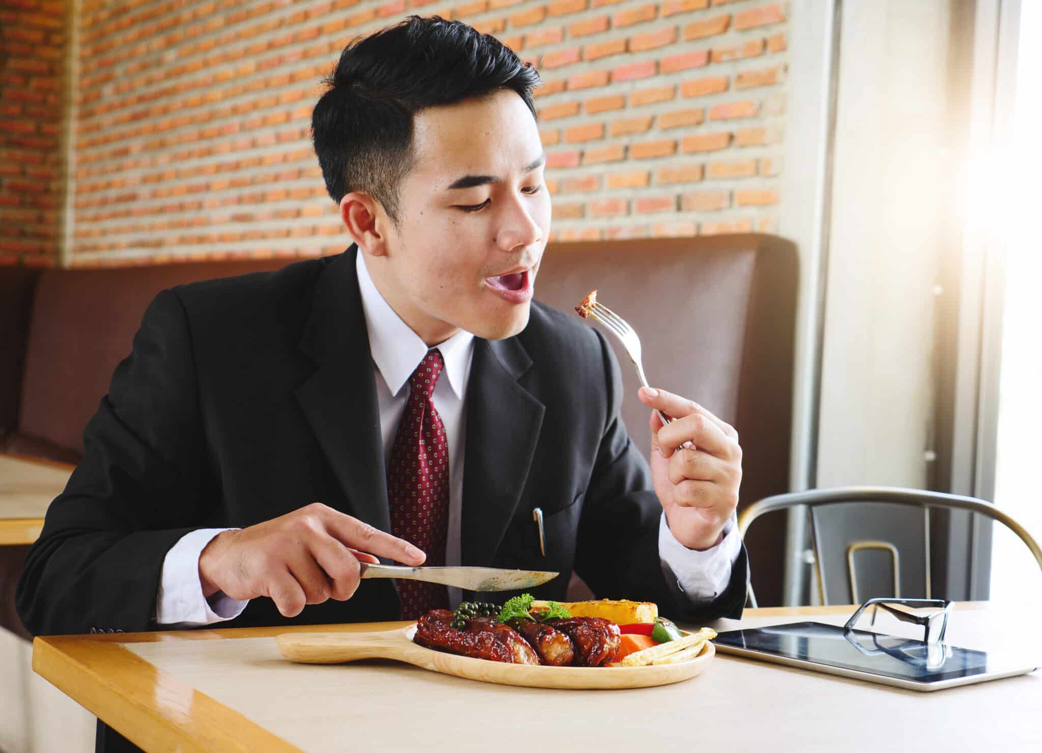 Egy öltönyös férfi ül egy étteremben, a villáján egy darab hús, a tányéron előtte hús és zöldségek