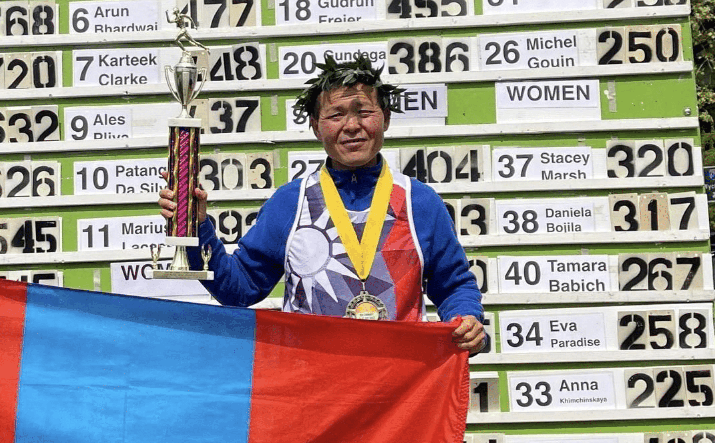 Budjargal Byambaa vegán ultramaraton futó az eredménytábla előtt, egyik kezével a kupáját fogja, másikkal a mongol zászlót