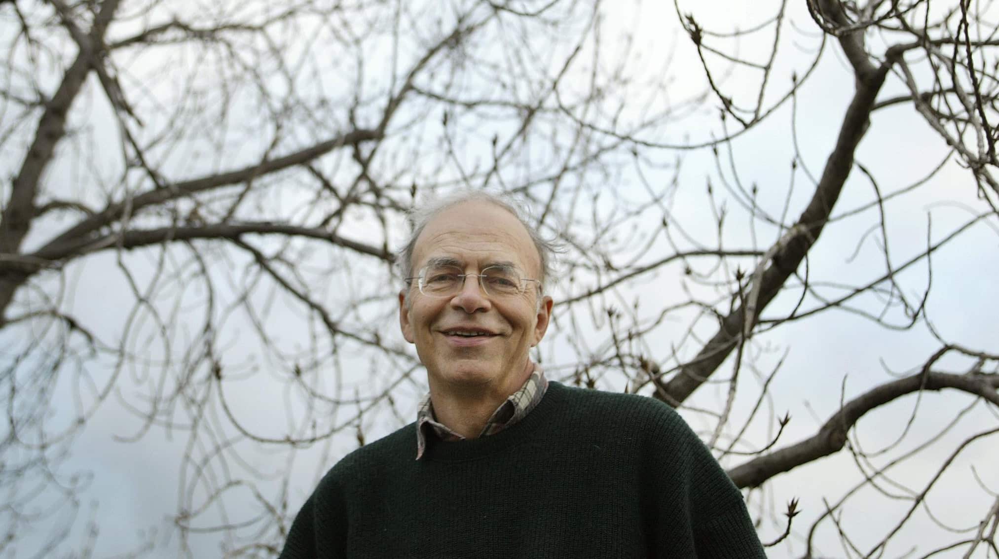 Peter Singer morálfilozófus mosolyog a kamerában, mellkastól felfelé látszik, kissé alulról fotózva, mögötte a szürkés ég és egy fa kopasz ágai