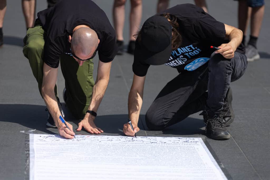 Földre kiterített papírt ír alá két férfi.