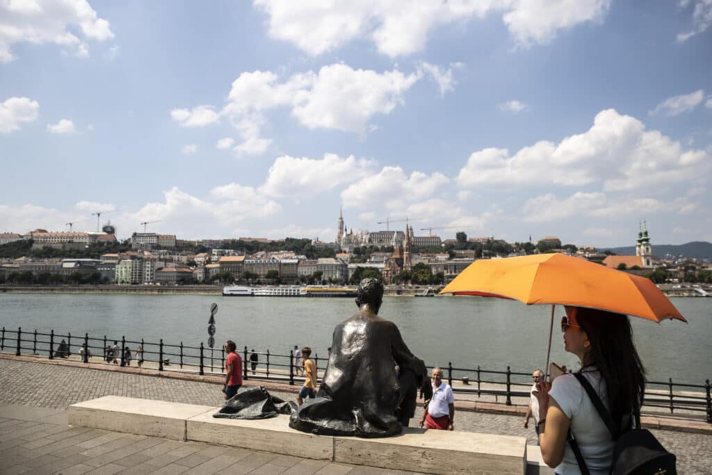 Budapesten, a Duna mentén sétál egy nő, narancssárga ernyővel az erős napsütés miatt. A folyó túloldalán látszik a budai oldal