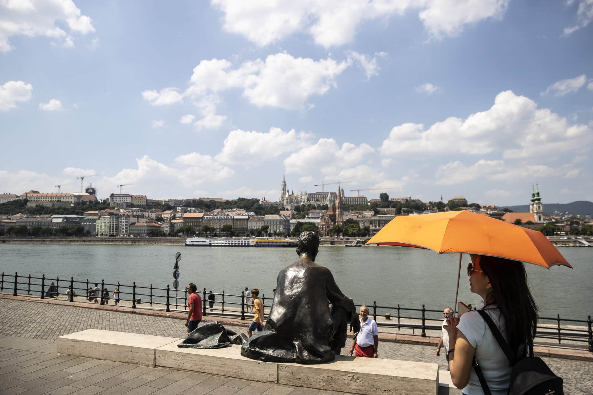 Budapesten, a Duna mentén sétál egy nő, narancssárga ernyővel az erős napsütés miatt. A folyó túloldalán látszik a budai oldal