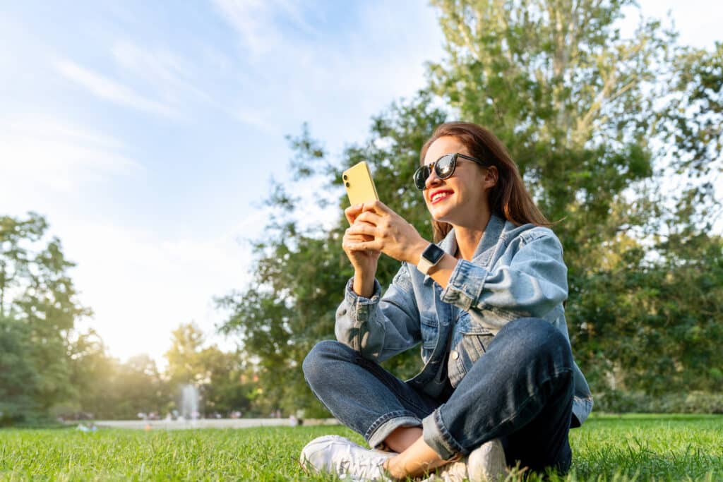 Napszemüveges nő farmernadrágban és farmerdzsekiben ül a füvön, a háttérben fák, és a telefonját nézi