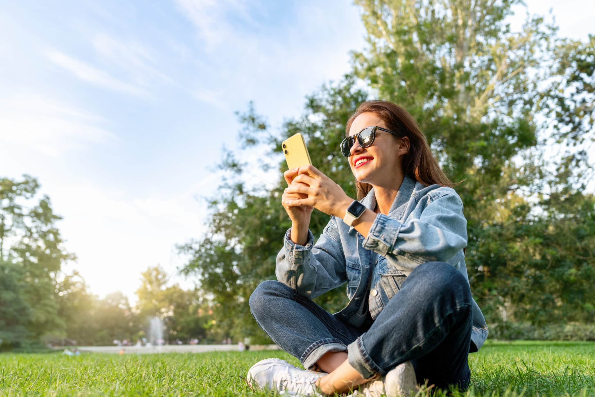Napszemüveges nő farmernadrágban és farmerdzsekiben ül a füvön, a háttérben fák, és a telefonját nézi