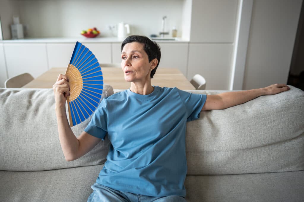 Nő kék pólóban ül a kanapén, kék legyezővel legyezi magát