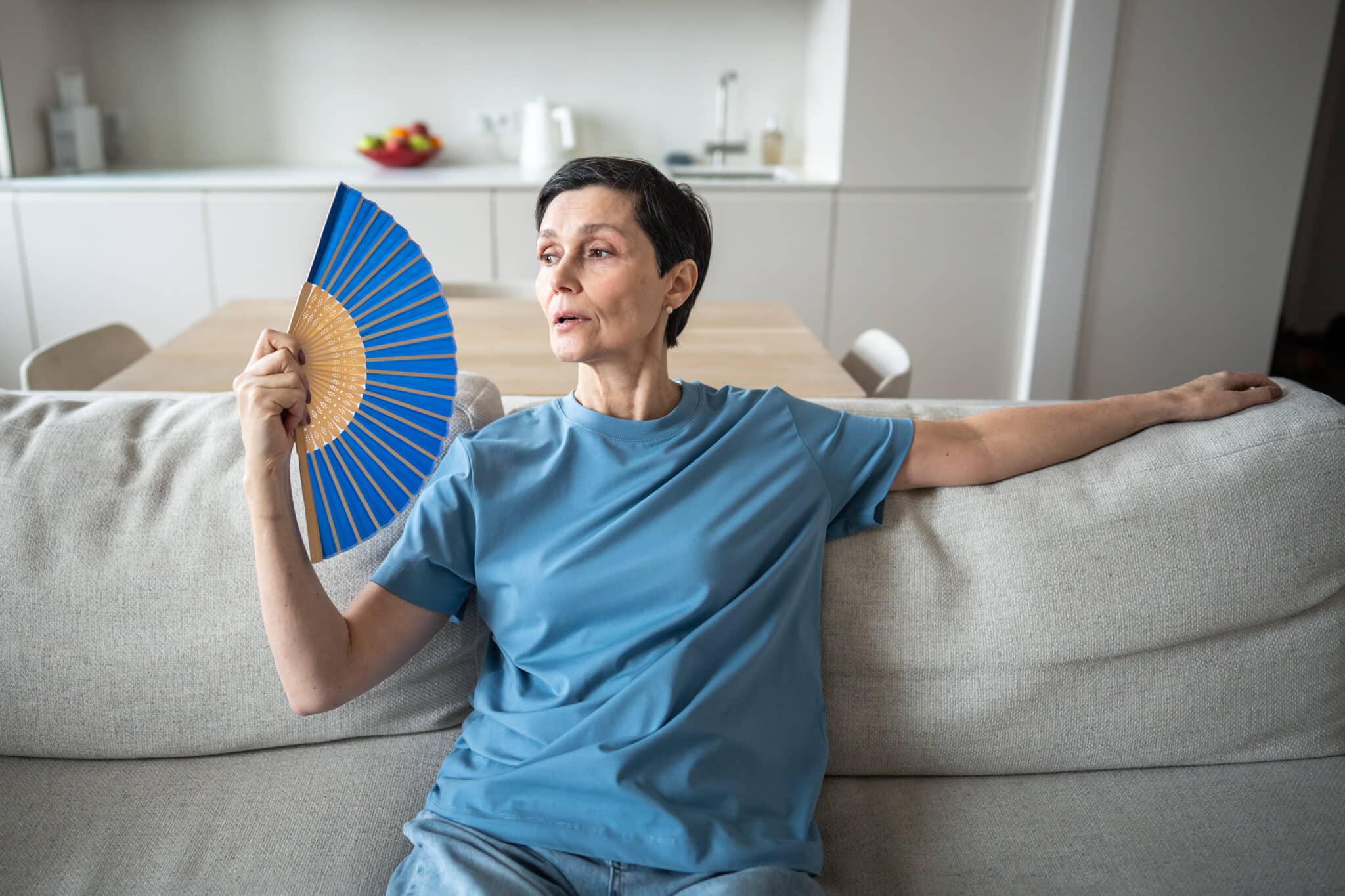 Nő kék pólóban ül a kanapén, kék legyezővel legyezi magát
