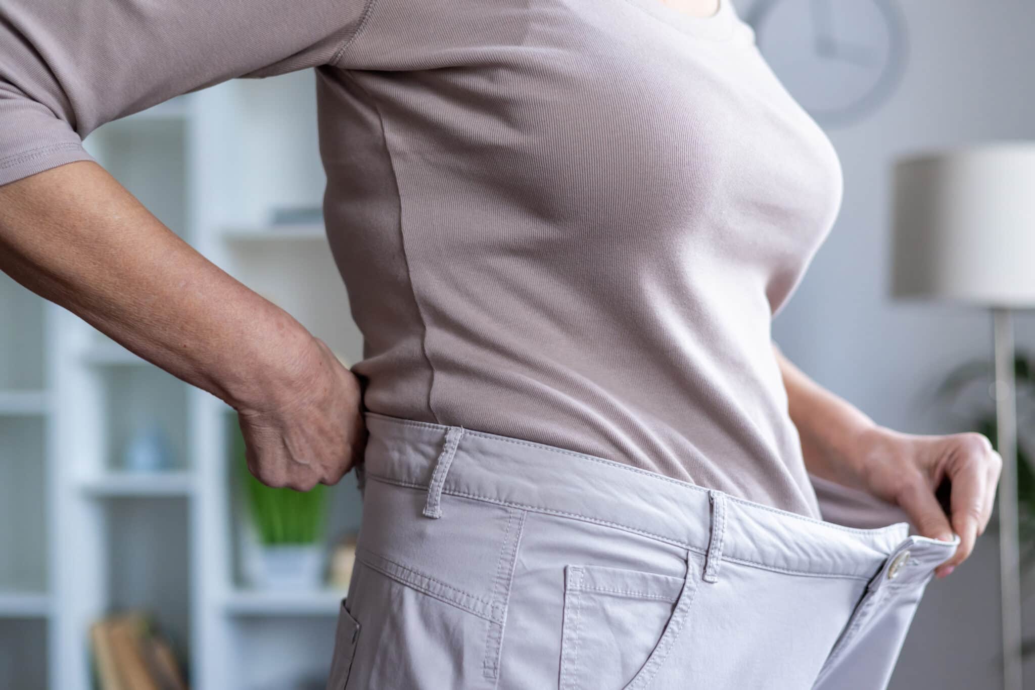 Drapp felsőt viselő nő mutatja egy nagyobb nadrágot tartva a derekánál, mennyit fogyott.