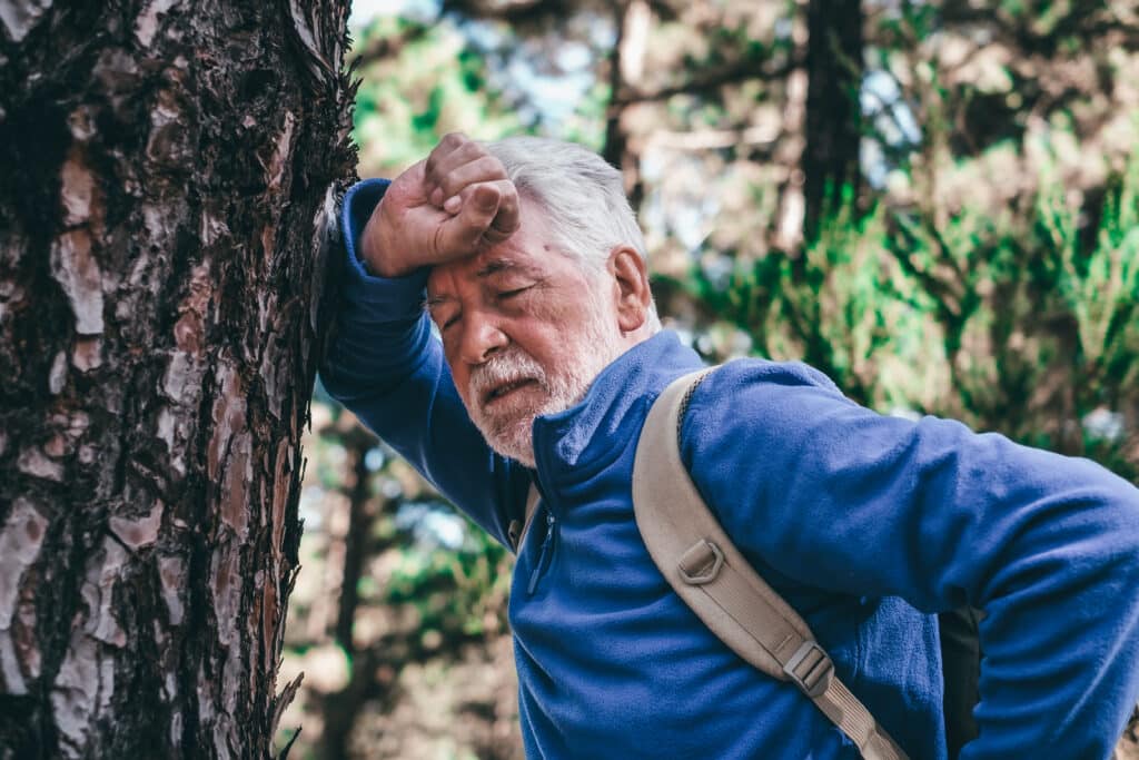 Idősödő férfi ősz hajjal és szakállal, kék pulóverben, hátizsákkal áll egy fának dőlve, kifulladva, szemét becsukja
