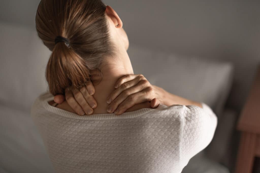 Copfos nő hátulról, mindkét kezével a nyaktövét, vállát fogja, jól láthatóan fájlalja