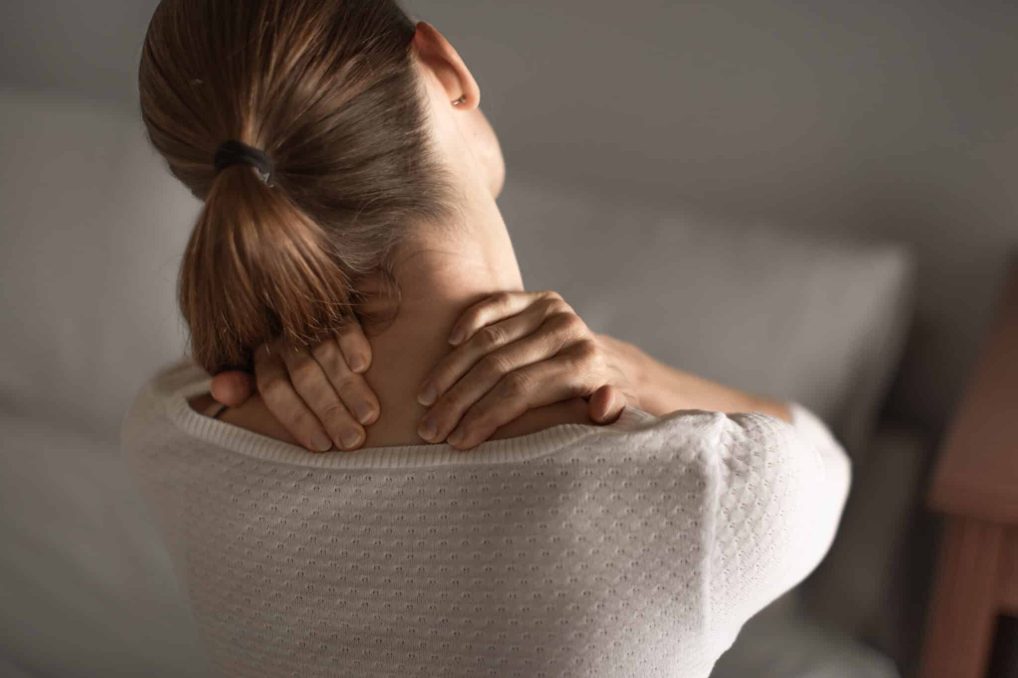 Copfos nő hátulról, mindkét kezével a nyaktövét, vállát fogja, jól láthatóan fájlalja