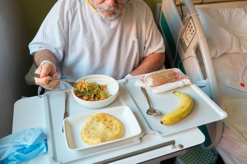 Idős férfi kórházi környezetben ül az ágy mellett, és tálcára rakott ételeket fogyaszt
